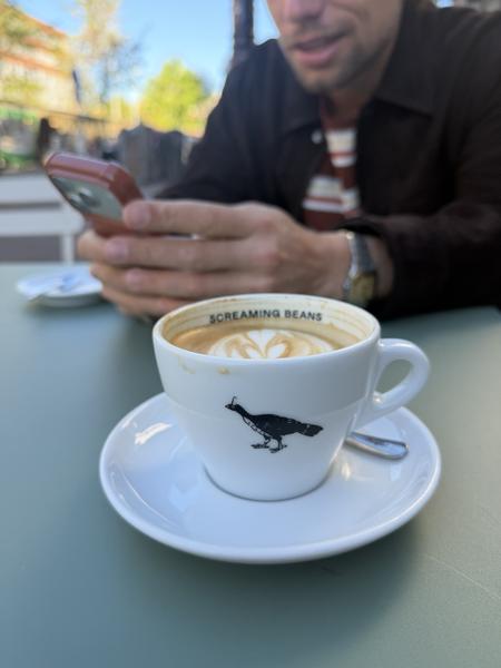 A close-up shot captures a latte with heart-shaped art on a light green table, with a person in the background engrossed in their smartphone